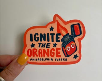 May include: A sticker with a cartoon design featuring a red lighter with flames and the text "IGNITE THE ORANGE PHILADELPHIA FLYERS" in blue and orange. The sticker has a peach-coloured border and star accents.