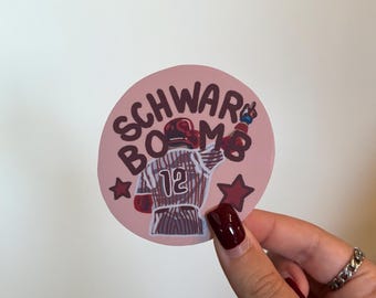 May include: A round sticker with the text "SCHWAR BOMB" in dark red lettering. The sticker features an illustration of a baseball player in a jersey with the number 12, along with red stars.