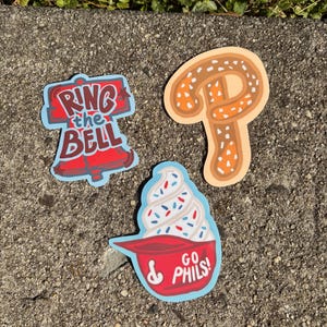 May include: Three colorful stickers on a gray surface. One sticker says "Ring the Bell" in red and blue. Another is a pretzel with brown and orange details. The third is a red bowl with a soft serve ice cream and the text "Go Phils!"