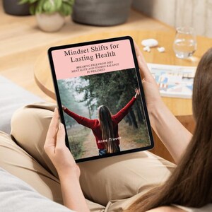 Könnte beinhalten: Ein Tablet zeigt das Buchcover von "Mindset Shifts for Lasting Health" von Anne Travel. Das Cover zeigt eine Frau mit ausgestreckten Armen in einem Wald. Das Tablet ist schwarz mit einem rosa Header, gehalten von einer Person.