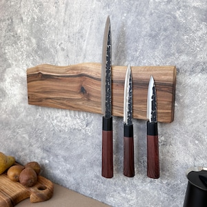 May include: A wooden knife holder with three knives. The knives have dark blades and brown handles. The holder is made of a light-colored wood and is mounted on a gray wall. The knives are of varying sizes.