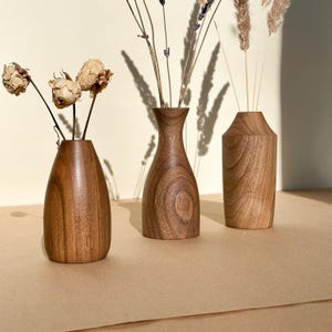 May include: Three wooden vases of varying shapes and sizes, each with a natural wood grain finish. The vases hold dried flowers and grasses, creating a rustic and natural aesthetic. The vases are displayed on a neutral-toned surface.
