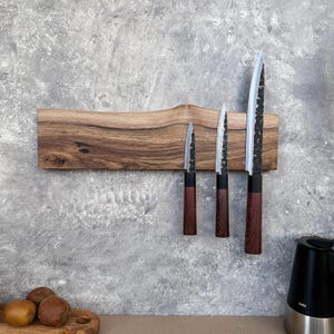 May include: A wooden knife holder with three knives. The holder is made of light brown wood with a natural edge. The knives have dark brown handles and silver blades. The blades have a textured appearance.