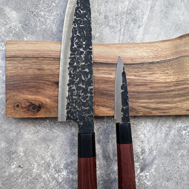 May include: Two Japanese-style kitchen knives with dark, textured blades and wooden handles. The knives are displayed on a wooden cutting board with a natural edge, set against a grey background. The blades have a unique, hammered appearance.
