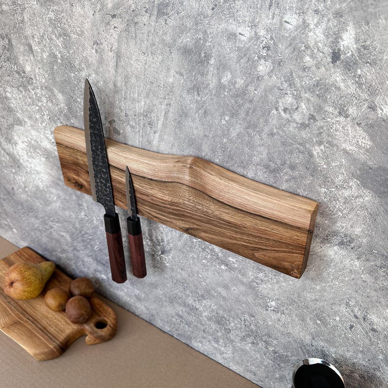 May include: A wooden magnetic knife holder with two knives attached. The holder is made of light brown wood and has a curved design. The knives have black blades and dark brown handles. A small wooden cutting board with fruit is in the foreground.