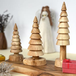 Könnte beinhalten: Drei Weihnachtsbaumdekorationen aus Holz in verschiedenen Größen. Die Bäume sind aus hellem Holz gefertigt und haben ein geschichtetes Design. Sie werden auf einem Holzbrett präsentiert, mit einer verschwommenen Engelsfigur im Hintergrund. Eine kleine rote Geschenkbox ist ebenfalls vorhanden.
