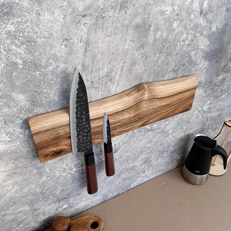 May include: A wooden knife holder with a natural edge, mounted on a grey wall, holds two knives. The larger knife has a textured, dark blade and a dark wood handle. The smaller knife has a similar design. The holder is approximately 40 cm long.