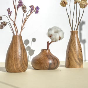 May include: Three wooden vases of different shapes and sizes, each with a different type of dried flower arrangement. The vases are made of dark brown wood with a natural grain pattern.
