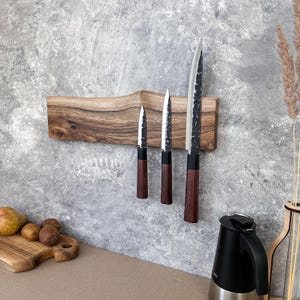 May include: A wooden knife holder with three knives. The knives have dark wooden handles and silver blades. The knife holder is made of a light brown wood and is mounted on a grey wall. The knives are of varying sizes.