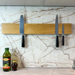 May include: A wooden magnetic knife rack with four knives attached to it. The rack is mounted on a white marble wall. A bottle of olive oil and a small jar are on a counter below the rack.