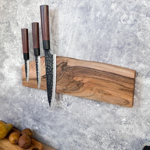 Puede incluir: Un soporte magnético de madera para cuchillos con tres cuchillos. Los cuchillos tienen hojas oscuras y mangos de madera. El soporte es de madera marrón claro con borde natural. Los cuchillos están dispuestos verticalmente.