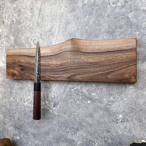 May include: A wooden magnetic knife holder with a single knife. The holder is made of dark brown wood with a wavy design. The knife has a dark brown handle and a silver blade. The knife is attached to the holder.