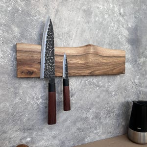 May include: A wooden knife holder with two knives. The knives have dark blades and reddish-brown handles. The wooden holder is rectangular with a curved design in the middle. The knives are of different sizes, one large and one small.