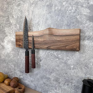 May include: A wooden magnetic knife holder with two knives. The holder is made of light brown wood with a natural edge. The knives have dark blades and dark brown handles. The holder is mounted on a grey wall.