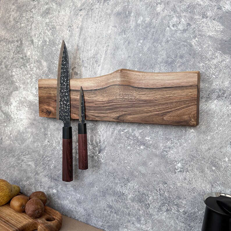May include: A wooden magnetic knife holder with two knives. The holder is made of light brown wood with a natural edge. The knives have dark blades and dark brown handles. The holder is mounted on a grey wall.