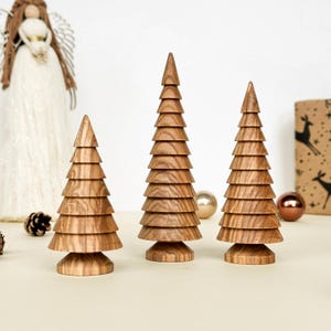May include: Three wooden Christmas tree ornaments in different sizes. The trees are crafted from light brown wood, featuring a layered design that creates a tiered appearance. The trees are placed on a light surface, with a decorative angel and baubles in the background.