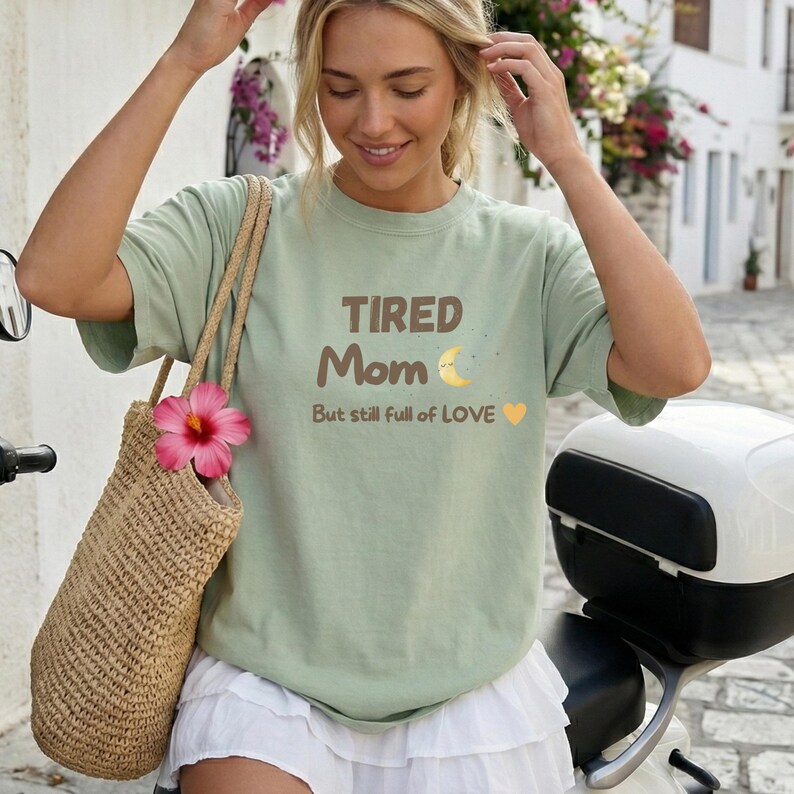 K&ouml;nnte beinhalten: Hellgr&uuml;nes T-Shirt mit dem Schriftzug "TIRED Mom" in Braun, einem Mond und dem Satz "But still full of LOVE" mit einem gelben Herz. Das Shirt wird mit einer Strohtasche und einer rosa Blume getragen.
