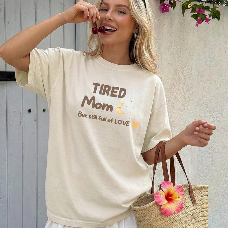 May include: Cream-colored t-shirt with the text "TIRED Mom But still full of LOVE" in brown and yellow. The shirt is paired with a straw tote bag adorned with a pink and yellow flower. The person is holding cherries.