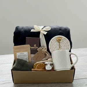 Op de afbeelding: Een cadeaupakket gevuld met een zwarte deken vastgebonden met een wit lint, een witte mok met verticale lijnen, een klein potje honing, thee, sokken en een onderzetter met een gouden lepel. De mand bevat een klein zakje en een doos chocolade.