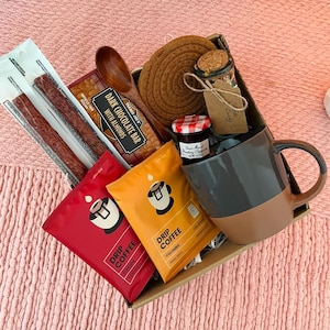 May include: A gift box filled with coffee, snacks, and a mug. Includes drip coffee packets, a dark chocolate bar with almonds, a jar of jam, a wooden spoon, and a brown and gray mug. The box sits on a pink surface.