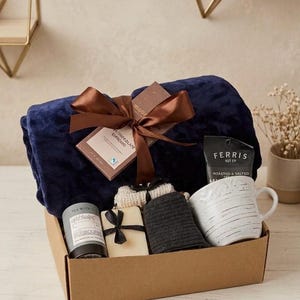 Op de afbeelding: Een geschenkdoos met een gevouwen marineblauwe pluche deken vastgebonden met een bruin lint, een chocoladereep, een zakje Ferris Nut Co. noten, een witte keramische mok met een textuur, een grijze kaars en een paar donkergrijze sokken.