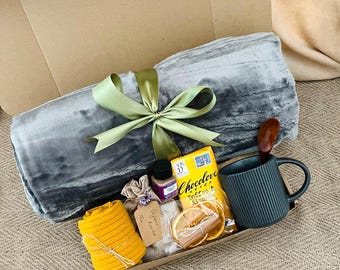 Può includere: Una confezione regalo con una coperta grigia legata con un nastro verde, un oggetto giallo arrotolato, una tazza con un cucchiaio di legno, una barretta di cioccolato e altri piccoli oggetti. La scatola è aperta, rivelando il contenuto.