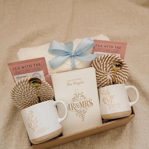 Può includere: Un cesto regalo contenente una coperta bianca legata con un nastro azzurro, due tazze bianche con disegni floreali, un diario con il testo "MR & MRS" e "365 Devotions For Couples", bustine di tè e due cucchiai dorati.