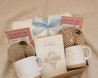 Puede incluir: Una cesta de regalo que contiene una manta blanca atada con una cinta azul claro, dos tazas blancas con diseños florales, un diario con el texto "MR & MRS" y "365 Devotions For Couples", sobres de té y dos cucharas doradas.