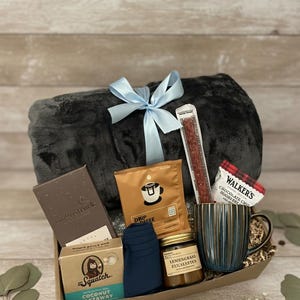 May include: A gift basket with a gray blanket tied with a light blue ribbon. The basket contains coffee, a mug, chocolate, a snack stick, socks, soap, and a jar of lemongrass eucalyptus. The mug is blue and brown.