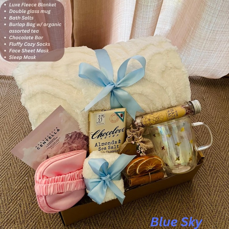 May include: A gift basket with a white fleece blanket tied with a light blue ribbon. It includes a double glass mug, chocolate bar, face sheet mask, and other self-care items. The text "Blue Sky" is at the bottom.