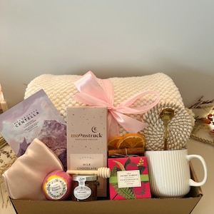 May include: A gift basket filled with self-care items. Includes a cream-colored blanket tied with a pink ribbon, a face mask, a sleep mask, a bath bomb, milk chocolate, honey, tea, a mug, and a spoon. The basket is in a brown cardboard box.