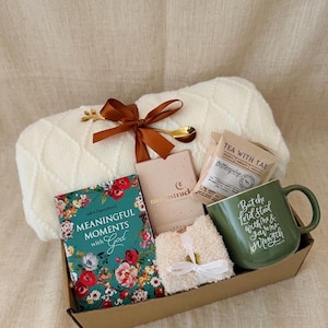 May include: A gift box containing a white cable-knit blanket tied with a brown ribbon and gold spoon, a book titled Meaningful Moments with God, a Moonstruck candle, a Tea with Tae packet, fuzzy white socks, and a green mug with white text.