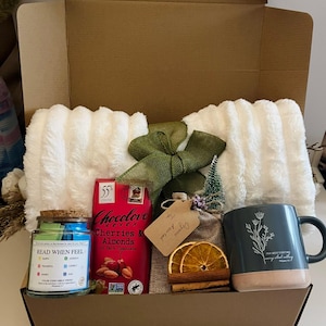 May include: A gift basket filled with a white fluffy blanket, a dark chocolate bar, a candle, a mug, and other items. The mug is dark blue with a floral design and the text "Pray about everything". The candle has the text "Read When Feel".