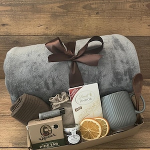 May include: A gift basket with a gray blanket tied with a brown ribbon. The basket contains a gray mug, wooden spoon, socks, lip balm, a box of pine tar soap, Lindt white chocolate, a jar of preserves, and dried orange slices.