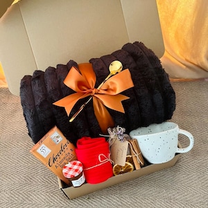 May include: A gift box containing a black plush blanket tied with an orange ribbon and a gold spoon. The box also includes a white speckled mug, chocolate, red socks, jam, and cinnamon sticks.