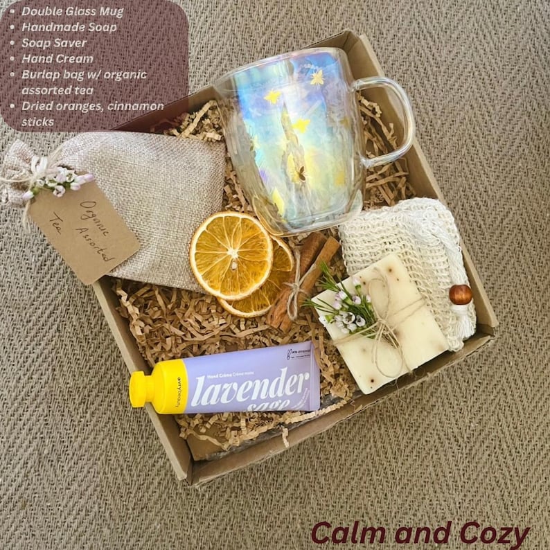 May include: A gift box with a double-walled glass mug, handmade soap, a soap saver, hand cream, a burlap bag of organic tea, dried oranges, and cinnamon sticks. The hand cream tube is lavender-scented. The words "Calm and Cozy" are at the bottom.