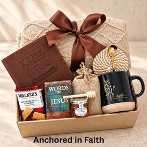 May include: A gift box containing a beige knit blanket with a brown ribbon, a brown journal embossed with "BE STRONG AND COURAGEOUS", a black mug with gold floral art and "be still", a small honey jar with a wooden dipper, and a "WORDS OF JESUS" book.