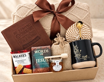 Puede incluir: Una caja de regalo con una manta de punto beige con un lazo marrón, un diario marrón grabado con "BE STRONG AND COURAGEOUS", una taza negra con arte floral dorado y "be still", un tarro pequeño de miel y un cucharón de madera para miel.