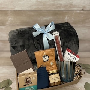 Op de afbeelding: Een cadeaupakket met een grijze deken vastgebonden met een lichtblauw lint. De mand bevat koffie, een mok, een snackstick, chocolade, zeep, sokken en een potje citroengras eucalyptus. De mok heeft blauwe en bruine strepen.