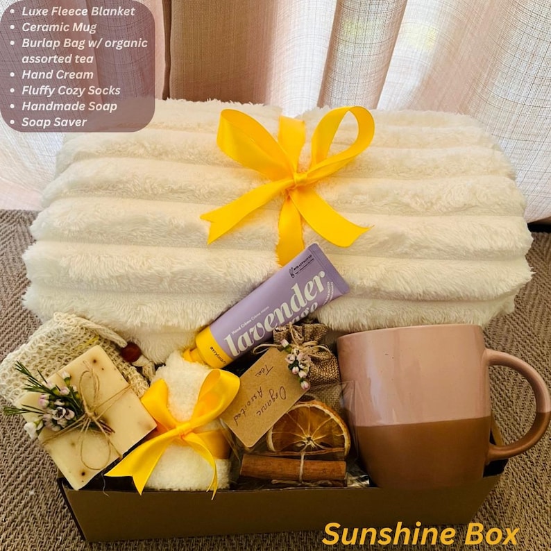 May include: A gift basket with a white fleece blanket, ceramic mug, hand cream, fluffy socks, handmade soap, and a soap saver. The basket is tied with a yellow ribbon and includes a burlap bag with organic tea. The text "Sunshine Box" is visible.