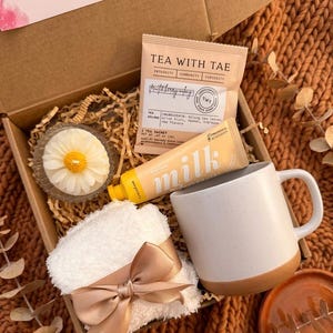 May include: A gift box containing a white speckled mug with a brown base, a tea sachet, a tube of "milk" lotion, a daisy-shaped candle, and a small white washcloth tied with a satin bow. The box is filled with crinkle paper.