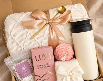 Può includere: Una confezione regalo con la scritta Comfort and Warmth contenente una coperta in maglia color crema con un cucchiaio dorato e un nastro di raso, un bicchiere nero e crema, una scatola rosa di cioccolata calda Lumi, una candela a fiore e sali.