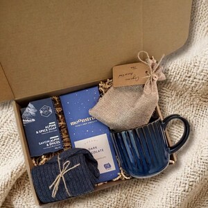 May include: A gift box containing a dark blue ribbed mug, dark chocolate, soap, socks, and a tea bag. The box includes the text "Alpine & Spice Soap" and "Moonstruck Portland Oregon". The tea bag is labelled "Organic Assorted Tea".