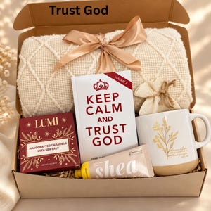 May include: A gift box containing a cream-colored knit blanket with a satin ribbon, a book titled Keep Calm and Trust God Volume 2, a box of Lumi handcrafted caramels, a tube of shea hand cream, and a white mug with gold floral print.