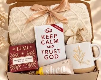 Puede incluir: Una caja de regalo que contiene una manta de punto color crema con cinta de satén, un libro titulado Keep Calm and Trust God Volume 2, una caja de caramelos artesanales Lumi, un tubo de crema de manos de karité y una taza blanca.