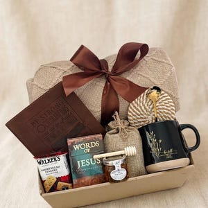 May include: A gift basket containing a beige cable-knit blanket tied with a brown ribbon, a brown leather journal embossed with text, a box of Walker's shortbread, a Words of Jesus book, a small honey jar with a dipper, and a black mug with gold floral art.
