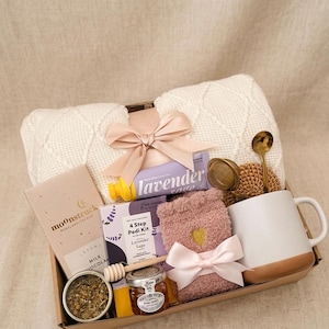 Op de afbeelding: Een cadeaupakket gevuld met zelfzorgartikelen. Inclusief een witte gebreide deken, lavendelzeep, een mok, honing, een chocoladereep, een voetverzorgingsset, zachte sokken en een thee-ei. Het pakket is vastgebonden met een roze strik.