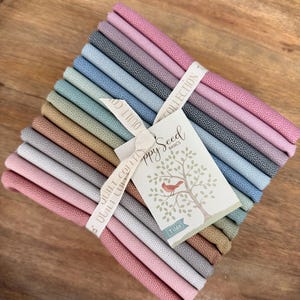 May include: A stack of fabric squares in various pastel colors, tied with a ribbon and a tag. The tag features a tree with a bird and the text "Happy Seed Basics" and "Tilda". The fabric has a dotted pattern.