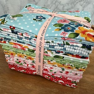 Puede incluir: Un montón de cuadrados de tela de algodón coloridos, atados con una cinta rosa. Las telas presentan diseños florales y de lunares en varios colores, incluyendo azul, rojo y verde. La cinta dice "Cotton sunshine and joy in every day."