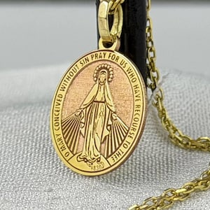 Puede incluir: Collar con colgante ovalado dorado con una imagen grabada de una figura religiosa. El colgante tiene la inscripción "O María concebida sin pecado, ruega por nosotros que recurrimos a ti". El collar es de cadena dorada.